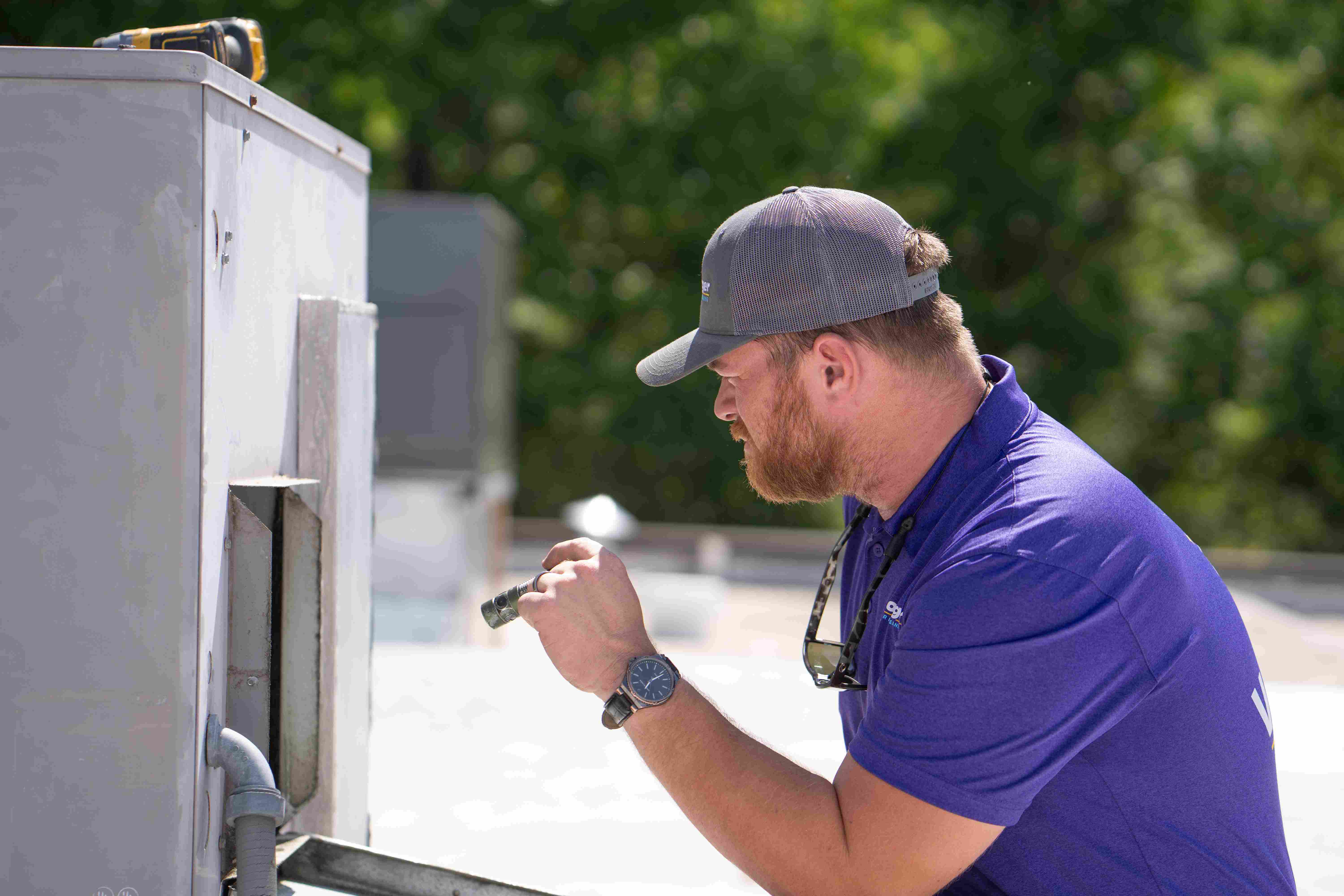 Voyager Air Duct Cleaning technician in a purple polo and gray cap inspecting a commercial rooftop HVAC unit with a tool in hand.